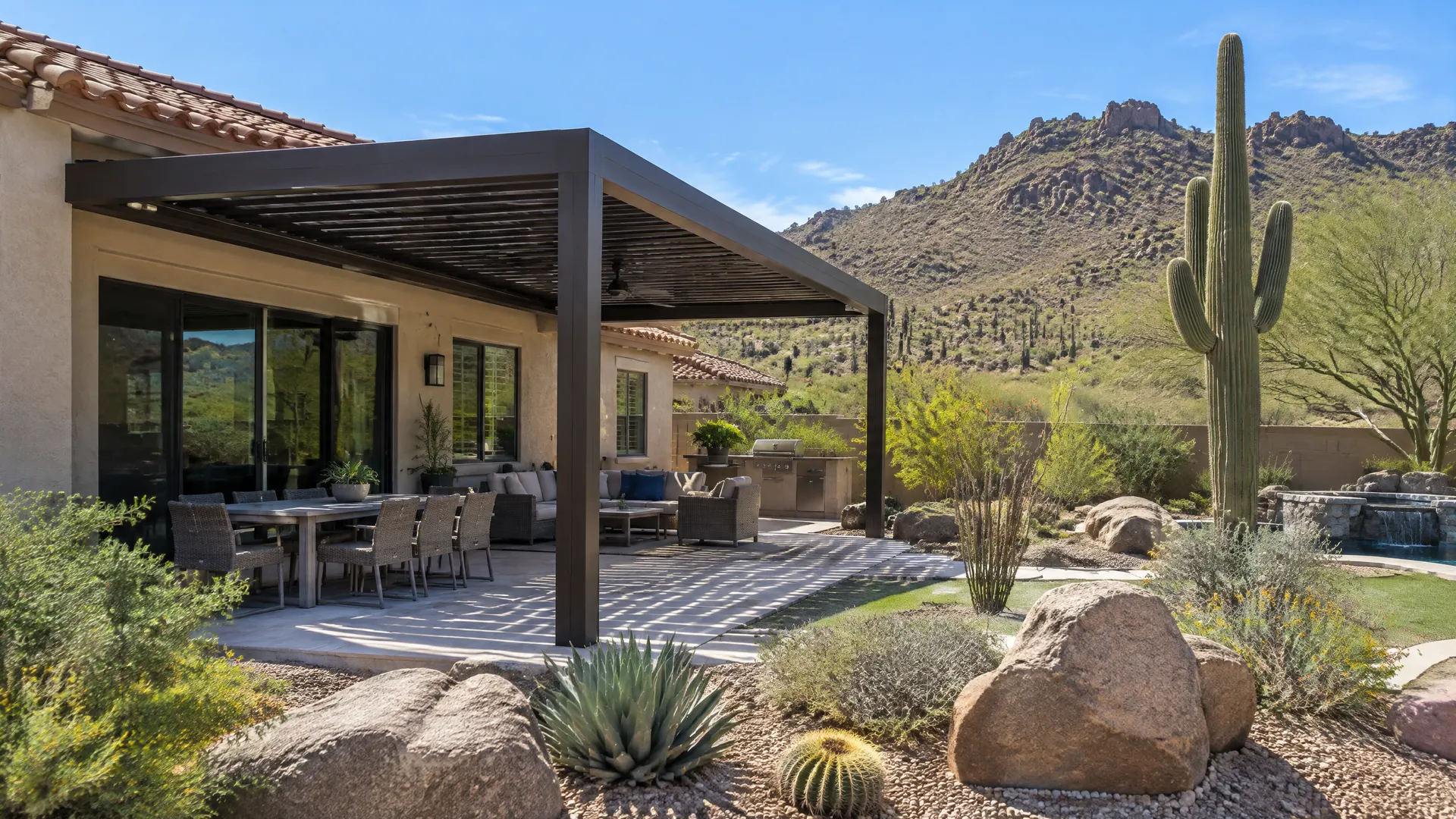 Backyard of a stucco home with a tile roof in far north Phoenix: dark bronze aluminum louvered pergola over a patio, natural desert landscaping with saguaro cactus and boulders, rocky desert hills behind