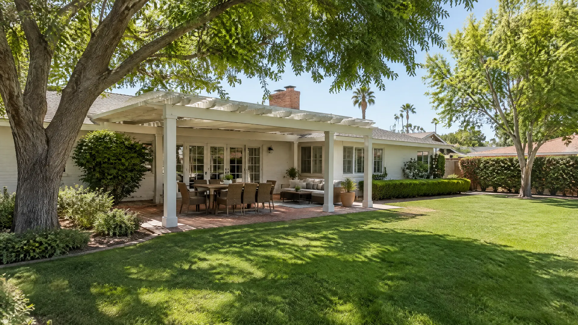 Backyard of a 1940s ranch-style home in a mature central Phoenix neighborhood: a painted white wood pergola over a brick patio, large shade trees, green lawn, low hedges