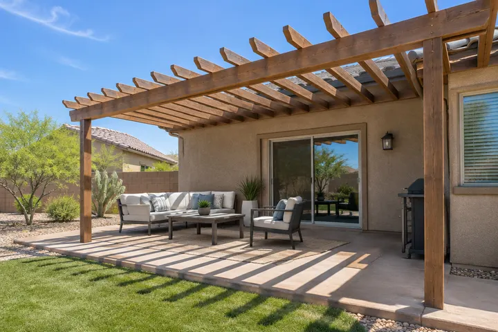 Open-roof wood pergola attached to the back wall of a single-story stucco Phoenix home over the patio off the sliding door, exposed rafters with blue sky visible between them and striped shadows below, no solid roof