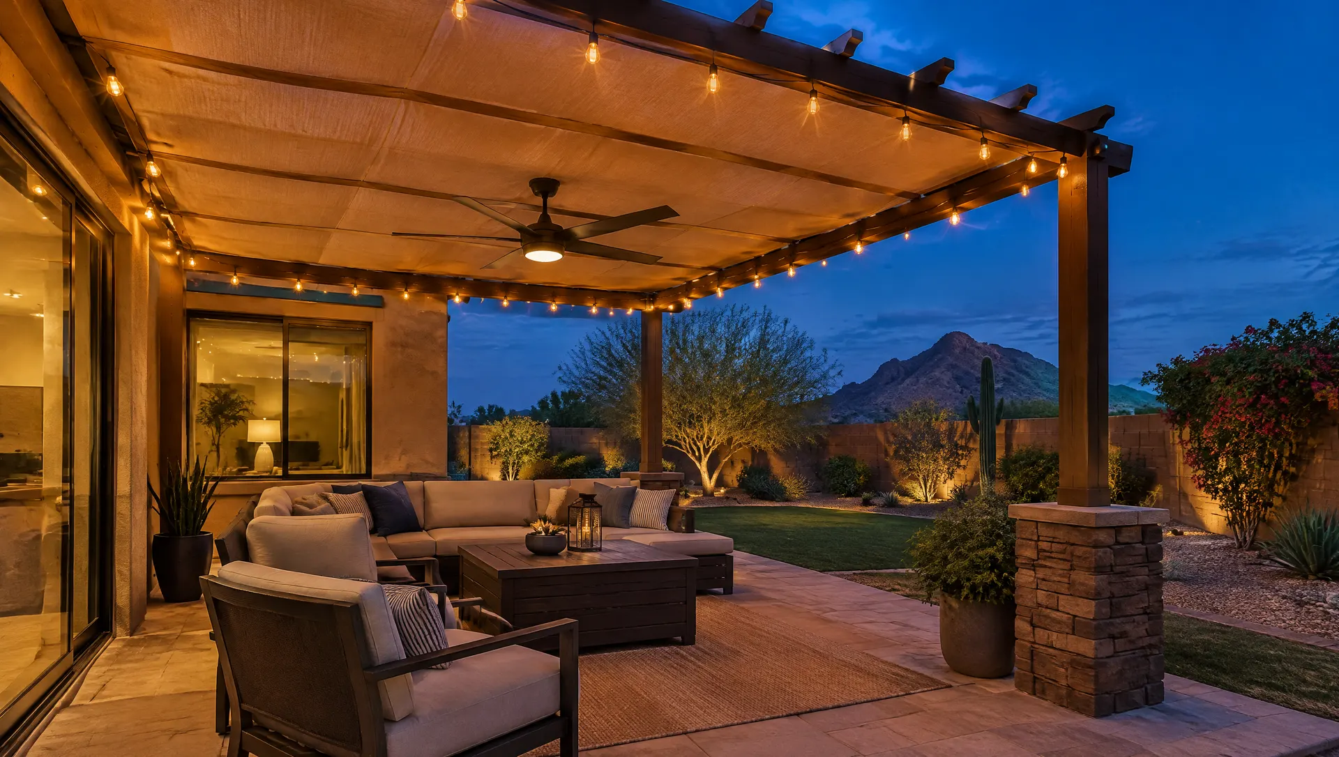 Pergola in a Phoenix backyard at dusk with a tan fabric shade canopy pulled across the rafters, warm string lights and an outdoor ceiling fan, cushioned seating, deep blue evening sky